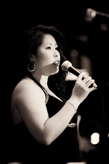 Anhthu singing into a microphone, lit against a dark stage
